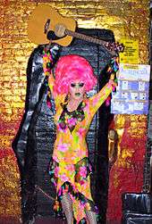 a drag queen, holding a guitar above her head. She has a bright pink wig on