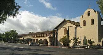 Photograph of Mission Santa Inés, showing the campanile on the right, the chapel at center, and the long, colonnaded walkway to the left.