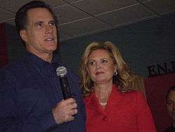 A tall, well-groomed pale-skinned man in his fifties with slightly greying dark hair wearing a dark blue shirt and holding a wireless microphone; to his side, a pale-skinned woman in her fifties with shoulder-length blond hair parted on the side, wearing red lipstick and a red jacket that almost obscures a double strand of white pearls around her neck; she is looking at him as he looks out at an unseen audience