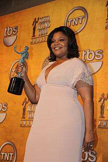 An African-American female with dark brown hair that reaches her shoulders. She is wearing a short sleeved light pink dress and a ring on her left hand. She is holding a greenish-blue statue that has a bronze plank with her right hand. In the background, there is an orange wall with logos and writing, such as the words "tbs" and "TNT" (which has a circle around it).