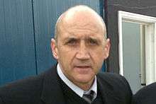 A middle-aged, balding man in a dark suit looks at the camera with a neutral expression on his face.