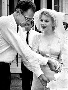 Cropped photo of Monroe and Miller cutting the cake at their wedding. Her veil is lifted from her face and he is wearing a white shirt with a dark tie.