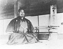A kneeling man in kimono in front of a sword stand