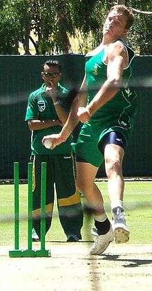 A man in the green South African cricket practice kit, in his bowling action. In the background are the wickets and another person.