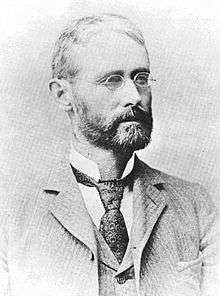 Head and shoulders of a white man with a full beard and wire-framed glasses, wearing a suit coat, vest, and tie.