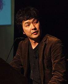 A Japanese man in a brown jacket and dark grey shirt standing at a podium