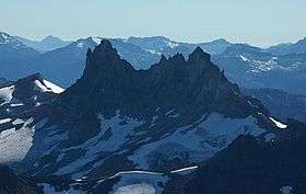 A dark rugged mountain rising over glacial ice in the foreground and glaciated mountains in the background.