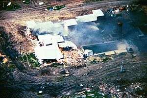 An aerial view from a helicopter of the Mount Carmel Center building. Large columns of smoke are arising from the left side of the building from a fire. One side of the building shows extensive damage. The building is surrounded by dirt paths.