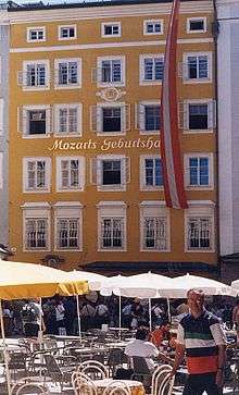  Facade of a tall brick building with rows of windows extending to five floors. Above the second row a sign indicates that this is Mozart's birthplace. In the foreground are sunshades and tables belonging to the modern café.