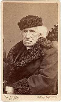 Photograph of seated, white-haired man in fur-trimmed coat and hat