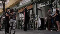 Nelly Furtado in a black jumpsuit walking on stilts along a city block.
