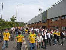 A group of people wearing predominantly yellow football shirts, walking along a road beside an association football stadium