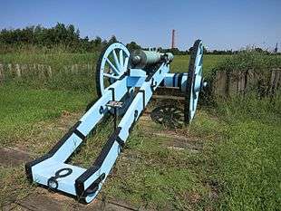 Photo shows a cannon on a carriage painted light blue and black. There is an open field in front of the gun. In the distance there is a tall smokestack with the lettering, "St Bernard Port".