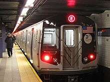 The back end of a subway train, with a red E on a LED display on the top. To the left of the train is a platform with a person walking away.
