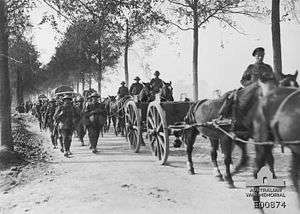 A black and white photograph of men in military uniforms marching on a road alongside carts being pulled by horses