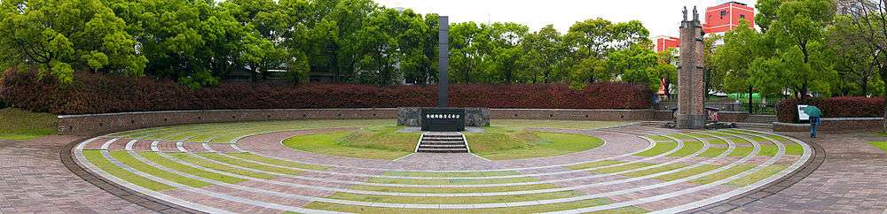 A rectangular column rises above a dark stone base with Japanese writing on it. It sits atop a grass mound with is surrounded by alternating circles of stone path and grass. The is a wall around the whole monument, and bushes beyond.