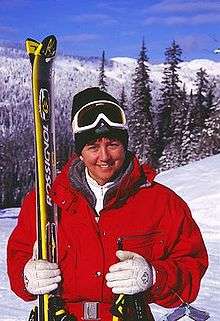 A woman smiles as she holds a pair of skis upright. She is wearing a thick red coat with white gloves and black toque.