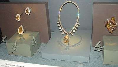 A museum display of jewelry items. Three brooches each consist of a large brown central gem surrounded by many clear small stones. A necklace has a large brown gem at its bottom and its string is all covered with small clear gems. A cluster-shaped decoration contains many brown gems.