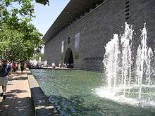 Daylight exterior of a long grey building with no windows and a single large semicurcular entrance arch, with a large rectangular shallow water feature in front of the building, including a fountain in the foreground. People are walking on a footpath next to the fountain and entering the building.
