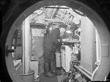 A picture, taken through a round hatchway, shows a machinery filled compartment. A single man, wearing a dark uniform, is bending over a table, lit by a desk-light.
