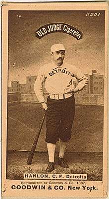 A baseball player is shown standing in his uniform, leaning on the end of a baseball bat.