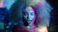 A portrait of a young woman with a blonde-styled afro in an orange top singing with her eyes closed as neon lighting shines upon her.