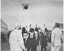 Female umpire and male umpire watch as young girls shoot a ball at a netball hoop.