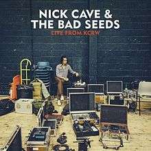 A man sits on a chair against a black stone wall amid cases of musical equipment. Above, white block text reads "Nick Cave & The Bad Seeds" and red block text reads "Live from KCRW".