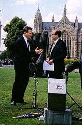 Two men in suits, stood on a grassy field in front of a Gothic style building. There is a tree on the left side, and microphone and recording equipment in the foreground on the floor.