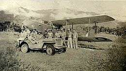 Military personnel and a jeep in front of a twin-engined biplane, with jungle and a mountain range in the background