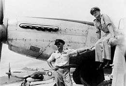 Two men in military uniforms and peaked caps, with a single-engined fighter plane