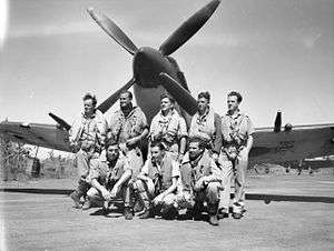 Black and white photograph of eight men wearing military uniforms posing in front of the nose of a single engined monoplane propeller fighter aircraft.