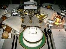 A set table with a white table cloth. There are many plates and glasses plus a menu visible on the table.