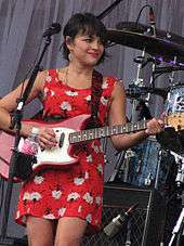 Smiling woman with short dark hair in red flower-print dress playing red and white electric guitar