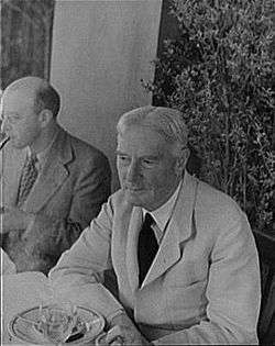 outdoor photograph of elderly man sitting at a table; he has white hair, and is clean shaven