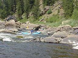 Canoers run rapids on a boulder-strewn river in the mountains.