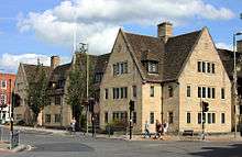 Adjoining buildings at the corner of a road, facing both ways; a mixture of gables and dormer windows at the roofline; built of light-coloured stone, they are two-storey in height below the roofs or gables