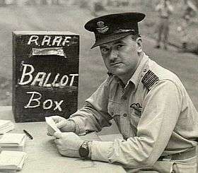 Man in military uniform with peaked cap seated at a desk beside a box marked "RAAF Ballot Box"