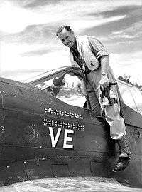 Full-length portrait of pilot smiling at the camera as he emerges from cockpit of single-engined monoplane with a number of black crosses and the letters "VE" prominently displayed on the fuselage