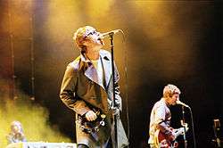 Two-thirds body shot of a singer wearing a coat with wide lapels; a guitar player is in the background. Both have short, blond hair.