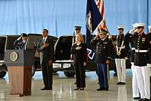 Obama and Clinton at a somber occasion, honoring the Benghazi attack victims at the Transfer of Remains Ceremony, held at Andrews Air Force Base on September 14, 2012. There are soldiers standing behind Obama and Clinton, and everyone is standing on a large wooden floor with their left hands to their side and their right hands on their upper chests.