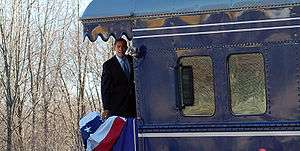 Obama is standing aboard the back platform of a train and looking to the side of the train. There is a red, white and blue banner hanging over the rear railing.