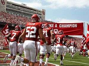 Football players run onto a football field in two rows under a crimson banner.