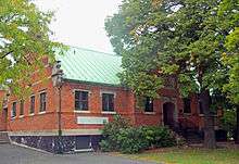 A one-story brick building with gabled roof and stepped parapet at the end and a large tree in front of its right side