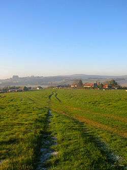 A lightly rutted field in winter, with several buildings in the middle distance and hills behind.  The buildings are partly obscured by fences, and consist of farm outbuildings, a large farmhouse with a red-tiled roof and chimneys, a smaller adjacent building in a similar style, and a two-storey house.