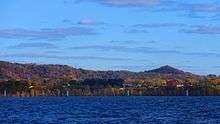 A landscape photo of Onalaska Wisconsin taken in the fall from Lake Onalaka shows a sunny sky of blue above with clouds and the cool blue waters of Lake Onalaska below in the foreground.  Sandwiched between is a horizontal ribbon that shows the city elevated 100 feet above the water and tree covered bluffs in the not to distant background.  A few houses along highway 35 are barely visible.