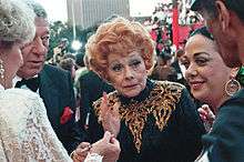 An aged Ball standing in a crowd of celebrities, wearing a black and gold sequinned dress with her characteristic red hair, looking fragile.