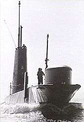 A submarine underway on the surface. A man is standing near the boat's bow sonar dome, while other personnel are visible on top of the sail