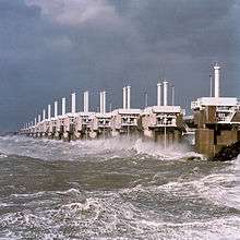 A long row of concrete towers with steel structures connecting them and a very rough sea