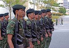 A row of young soldiers wearing green camouflage uniforms and dark green berets and holding rifles, standing at attention.